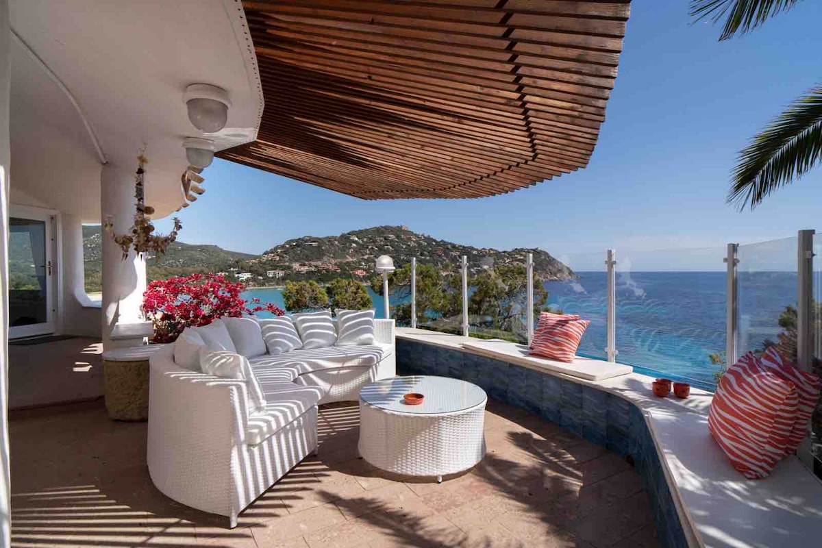 A shaded terrace features comfortable seating arranged around a round table. A striking view of the coastline and turquoise sea is visible, framed by lush greenery and colorful flowers. The overhead wooden structure provides protection from the sun, creating a serene outdoor space.