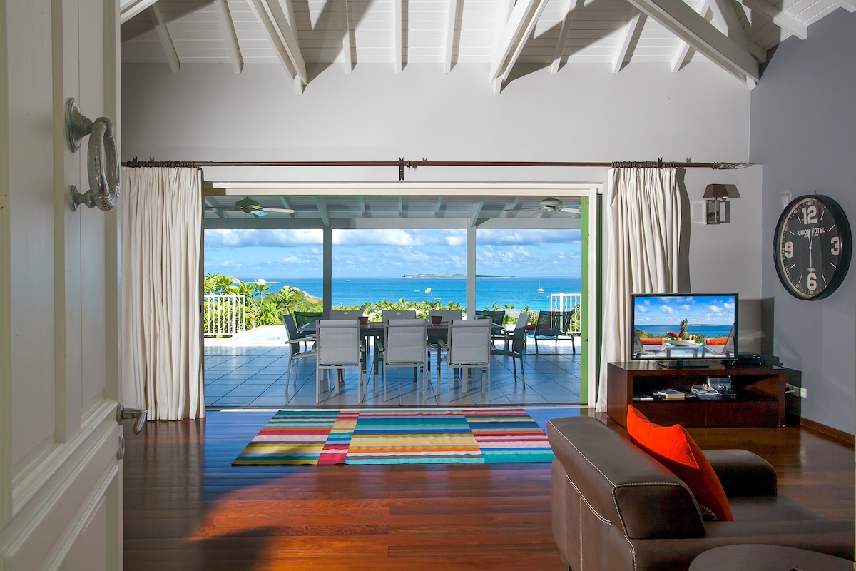 A spacious indoor living area is presented with large glass doors opening to a terrace with panoramic ocean views. A colorful striped rug lies on polished wooden floors, while a flat-screen TV and a classic clock provide warmth and functionality to the room.