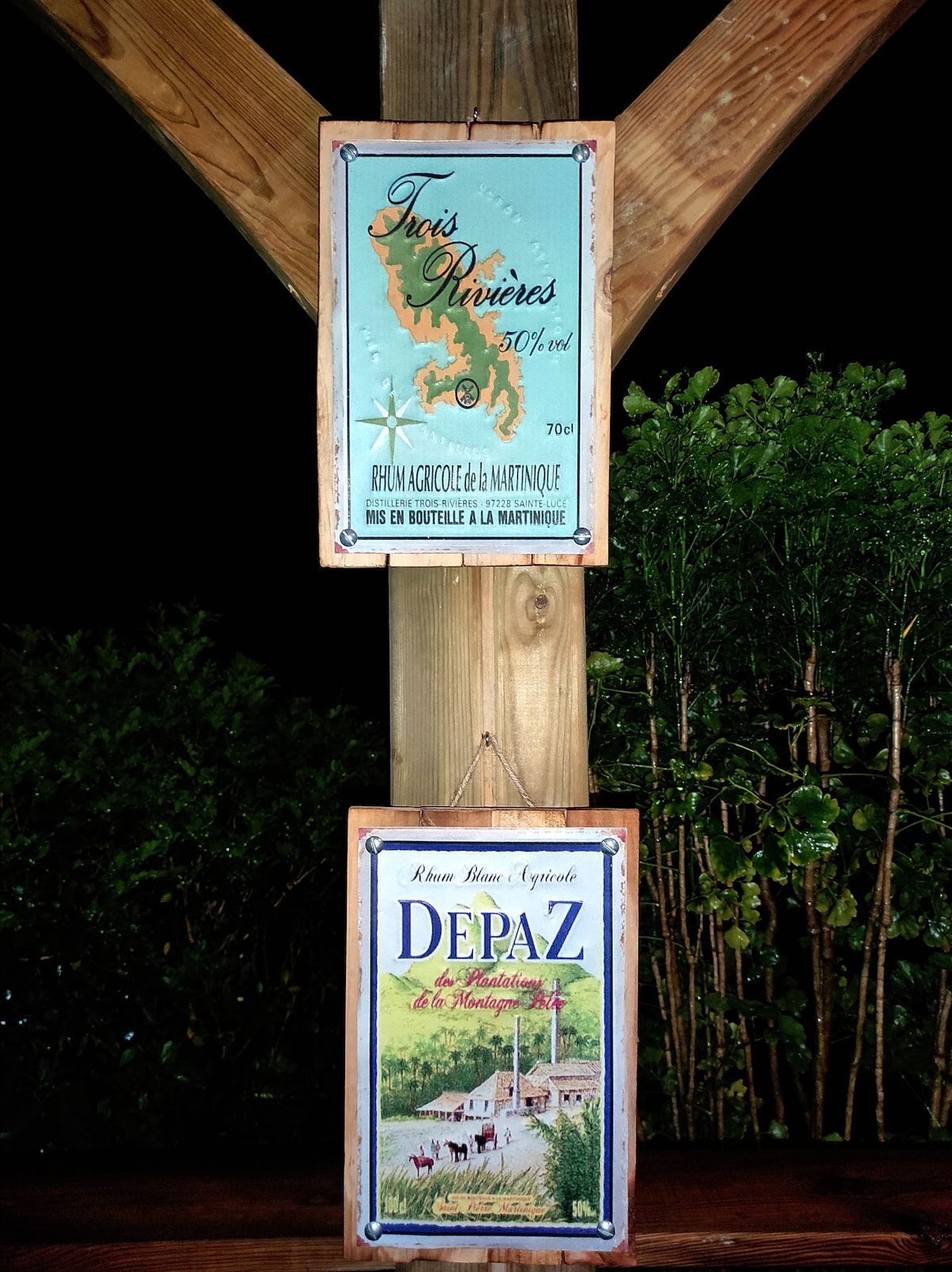 Two wooden signs are displayed under ambient lighting. The top sign features a map of Trois Rivières, while the bottom sign showcases a vintage-style advertisement for Depaz rum. The natural surroundings include greenery, enhancing the rustic atmosphere.
