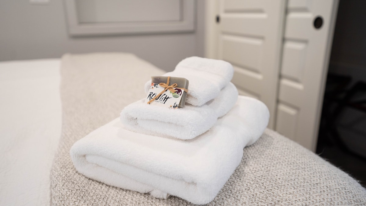 A stack of neatly folded white towels is placed on a light beige throw blanket on the bed. A small decorative tag is tied with twine, adding a thoughtful touch to the presentation. Soft gray walls and a partially visible door provide a calming backdrop.