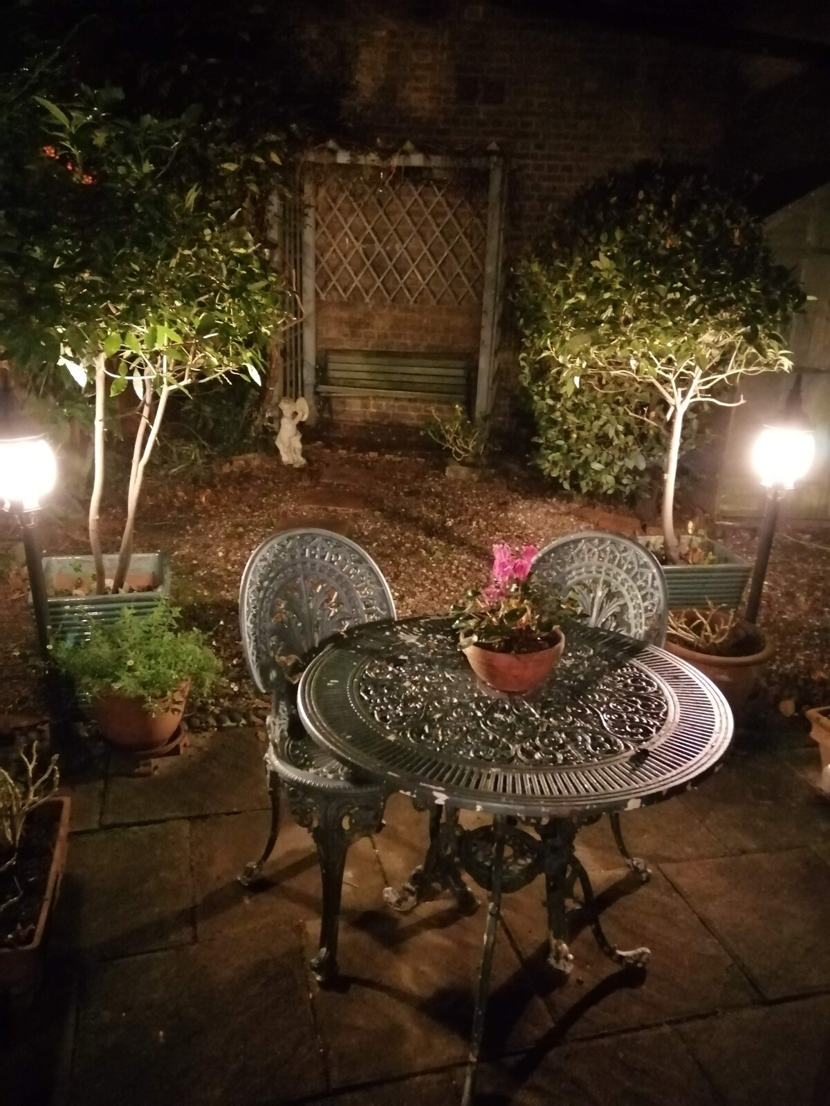 A charming garden space features a decorative metal table surrounded by two intricately designed chairs. Soft lighting illuminates the area, highlighting the greenery and potted plants. In the background, a statue is partially visible behind the garden’s foliage.
