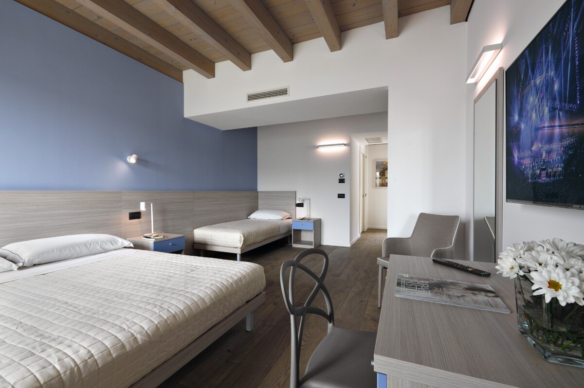 A well-lit room features two beds positioned against a light wood wall with a soft blue accent. A small desk with a chair sits by the window, while artwork and modern fixtures enhance the space, providing a comfortable environment for relaxation.