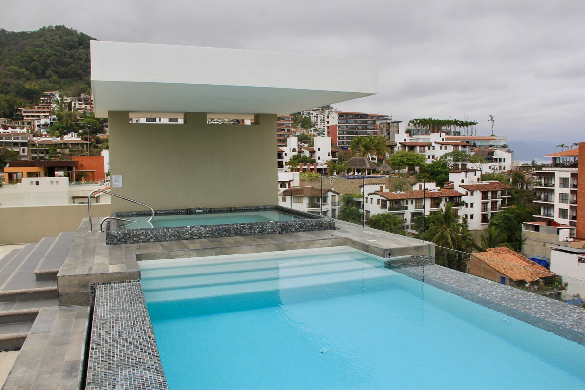 A rooftop pool area is presented, featuring a shimmering infinity pool that transitions to a hot tub. The surrounding landscape includes modern buildings and lush greenery, with a scenic backdrop of hills. The area is designed for relaxation, offering unobstructed views of the neighborhood.