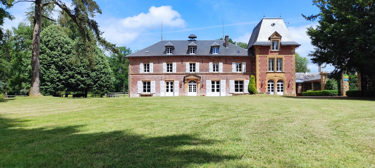 The exterior of a historic château is showcased, highlighting its red-brick façade and distinctive architecture. Lush green lawns and surrounding trees provide a natural backdrop. Large windows can be seen, contributing to the charm of the property set in a serene landscape.