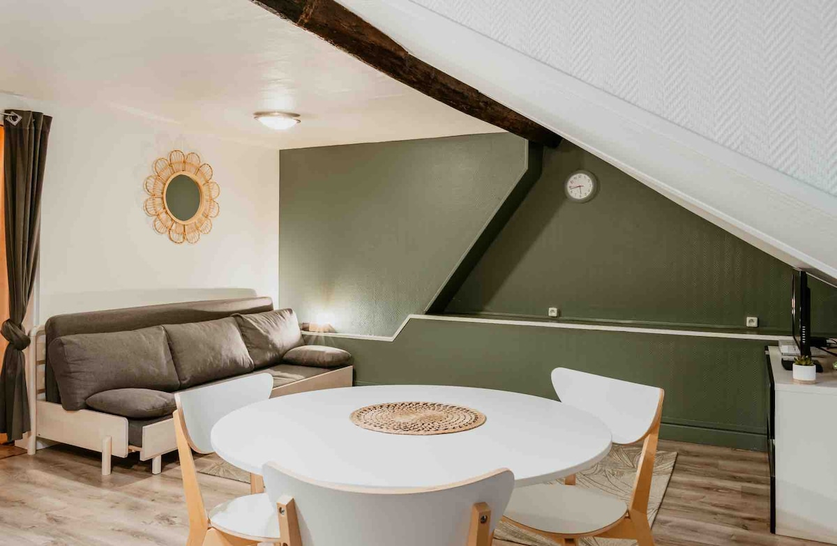 A cozy living area features a gray sofa bed and a circular dining table surrounded by white chairs. Soft lighting from a ceiling fixture and a round mirror enhance the inviting space, while a wall clock adds a functional touch.