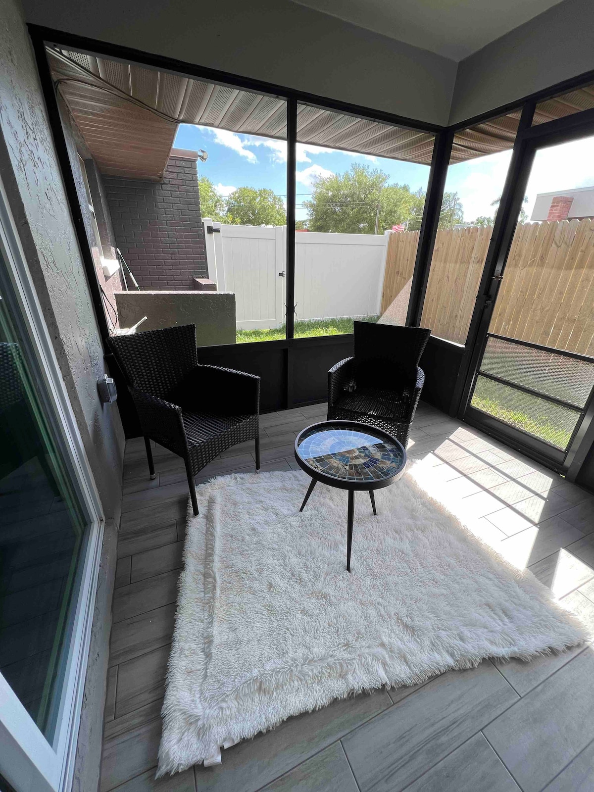 A screened-in porch features two black wicker chairs positioned beside a small round table. A light-colored area rug is laid on the floor, and natural light illuminates the space through the screens. A green lawn is visible outside.