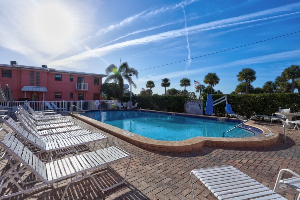 A heated swimming pool is surrounded by lounge chairs, set against a backdrop of palm trees and sunny skies. The pool area is bordered by a brick patio, offering a relaxing space for guests to enjoy the outdoors.