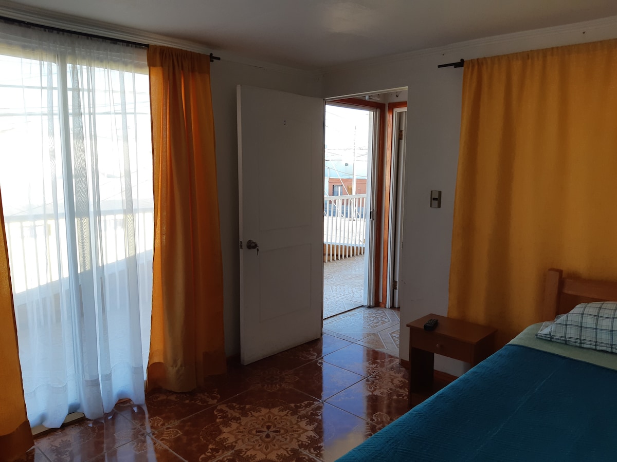 A bright room features large windows dressed in sheer and solid yellow curtains, allowing natural light to fill the space. A wooden door leads to an outdoor area, while a small bedside table and a neatly made bed contribute to the inviting ambiance.