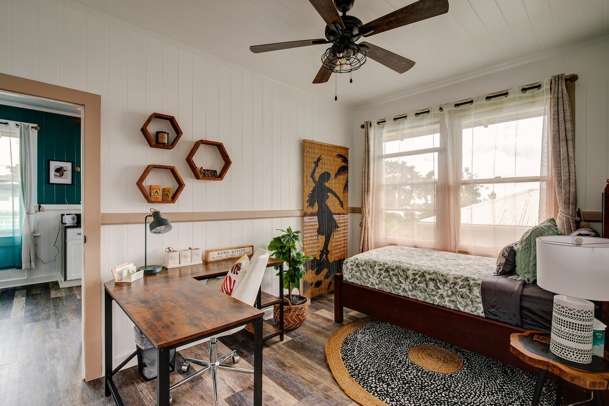A well-lit bedroom features a spacious bed with decorative bedding, complemented by a ceiling fan overhead. A rustic wooden desk and chair are positioned beside the bed. Natural light filters through large windows, partially covered by light curtains. A decorative plant adds a refreshing touch.