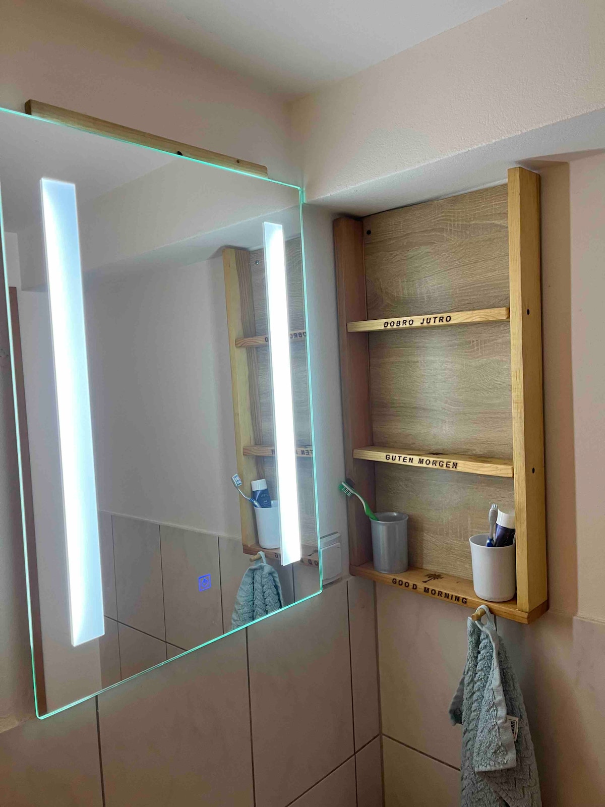 A well-lit bathroom features a modern mirror with integrated LED lights. Wooden shelves display toiletries, while a soft towel hangs nearby. Neutral tiles and subtle color choices create a clean, uncluttered space.