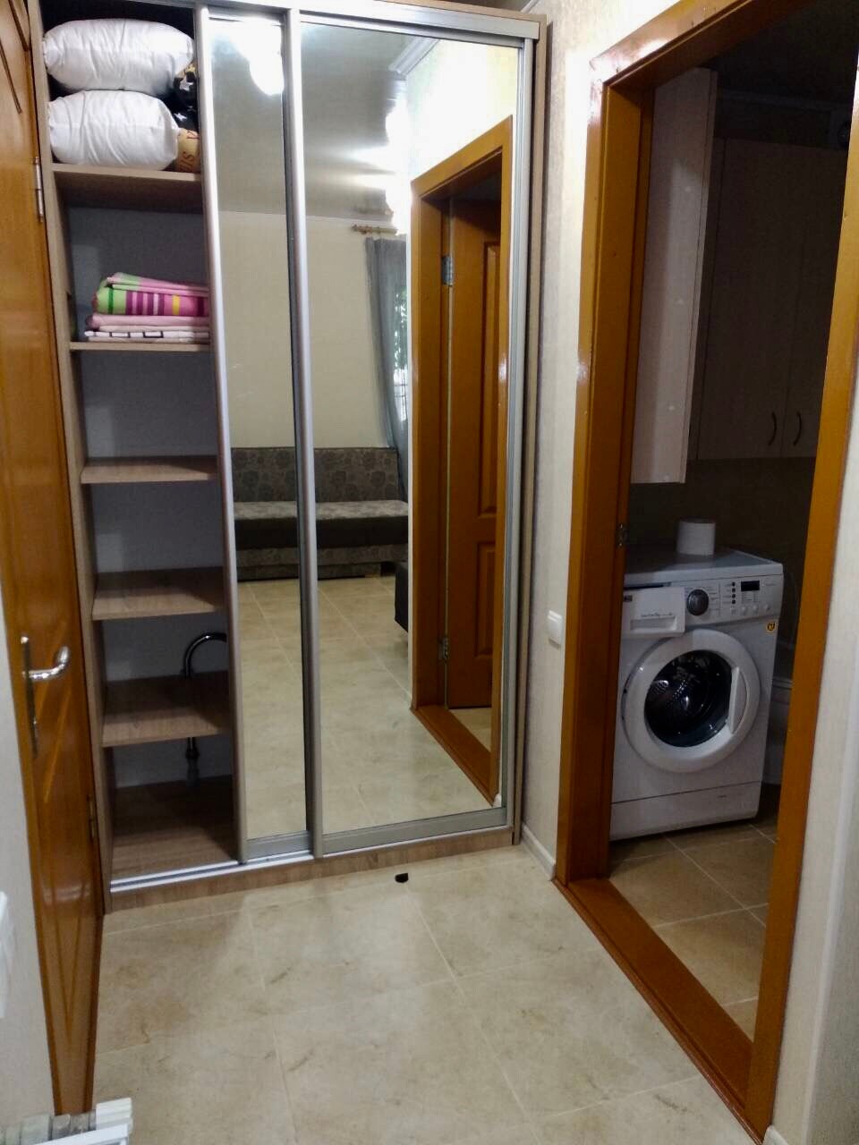 A functional entryway showcases mirrored sliding doors and an open shelving unit. To the right, a washing machine is positioned next to a door leading to the main living area. A comfortable seating area is visible in the background, adding an inviting touch to the space.