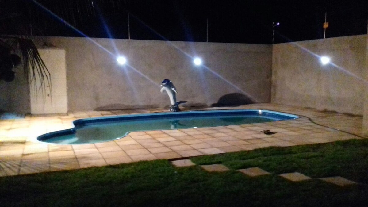 A rectangular pool is illuminated by soft lighting, casting a calm glow over the surrounding patio area. An artistic dolphin sculpture is positioned at the shallow end of the pool, while neatly trimmed grass borders the tiled surface.