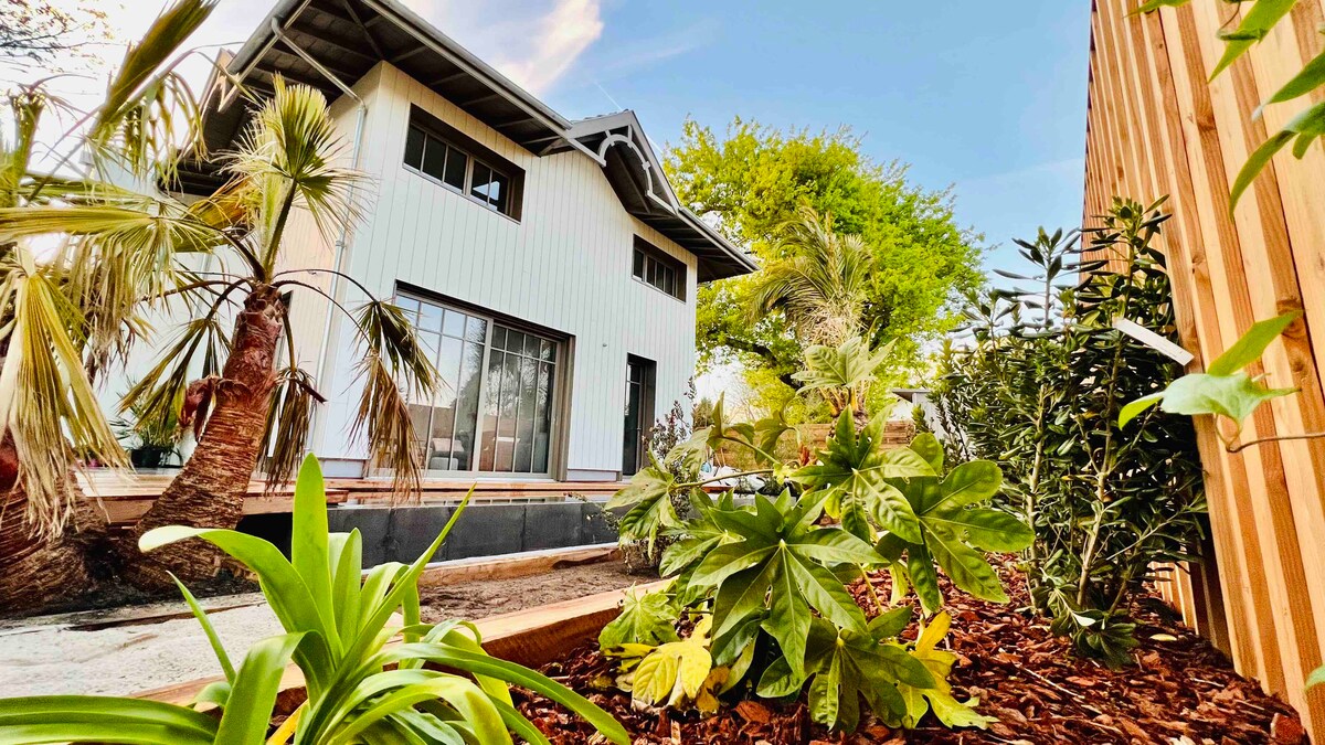 A modern wooden structure is framed by lush greenery, including various plants and palm trees. Large windows suggest ample natural light, while a wooden deck provides a welcoming entrance. The garden features decorative mulch that enhances the outdoor space.