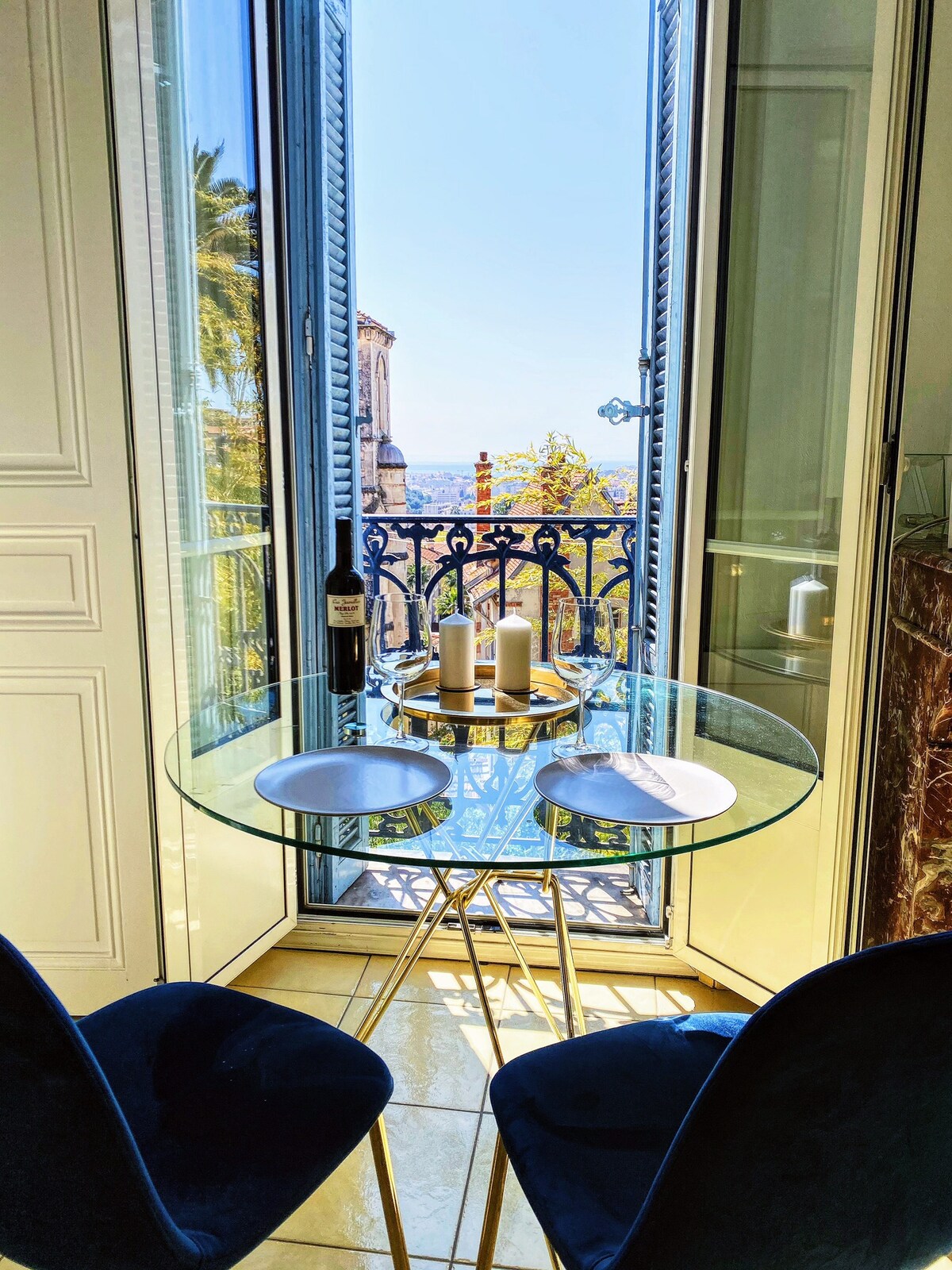 A small dining area features a round glass table set for two, with plates and candles arranged at the center. Two blue chairs are positioned around the table. A balcony door opens to reveal a scenic view, allowing natural light to enhance the space.