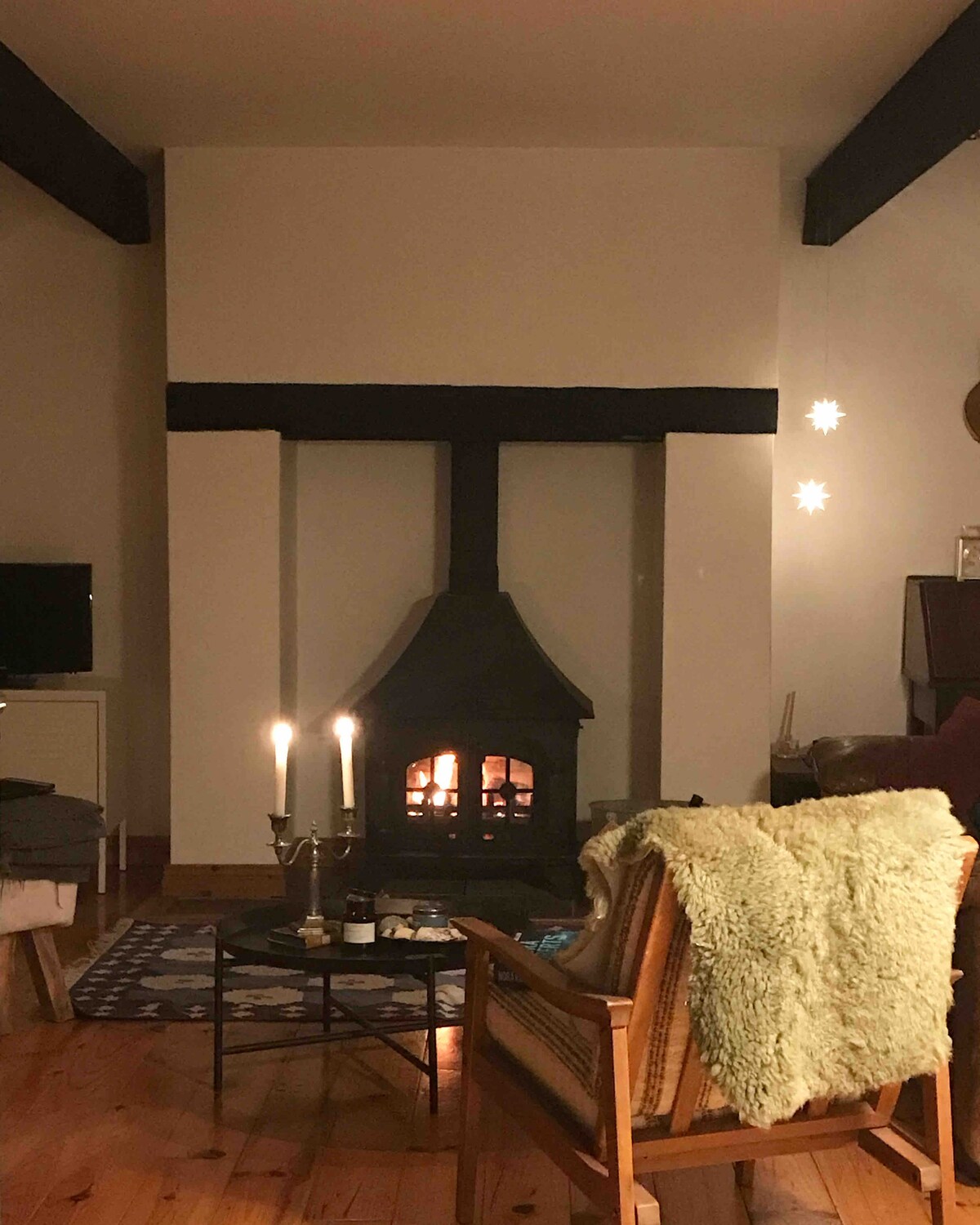 A cozy living area is highlighted by a wood-burning stove against a neutral wall. A woven chair with a soft throw is positioned nearby, with a coffee table displaying candles. The warm glow from the stove enhances the inviting setting, creating a relaxing atmosphere.