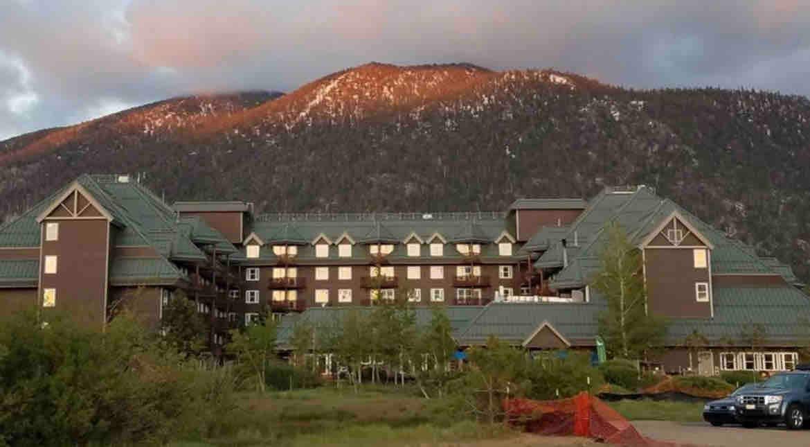 The exterior of a large, multi-story building is showcased against a backdrop of mountains. The structure features a green and brown color scheme, with multiple balconies visible on the upper levels. Surrounding greenery and natural landscape are present in the foreground.