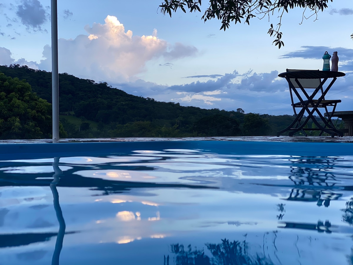 A serene view of a calm pool reflects the surrounding landscape under a softly lit sky. In the foreground, a wooden table and two bottles can be seen near the pool's edge, inviting relaxation with nature in the backdrop.