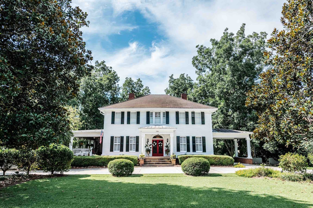 A historic two-story home is framed by lush greenery and vibrant trees. The white exterior is complemented by dark green shutters and a welcoming red front door. A spacious porch extends along the front, inviting guests to enjoy the outdoor surroundings.