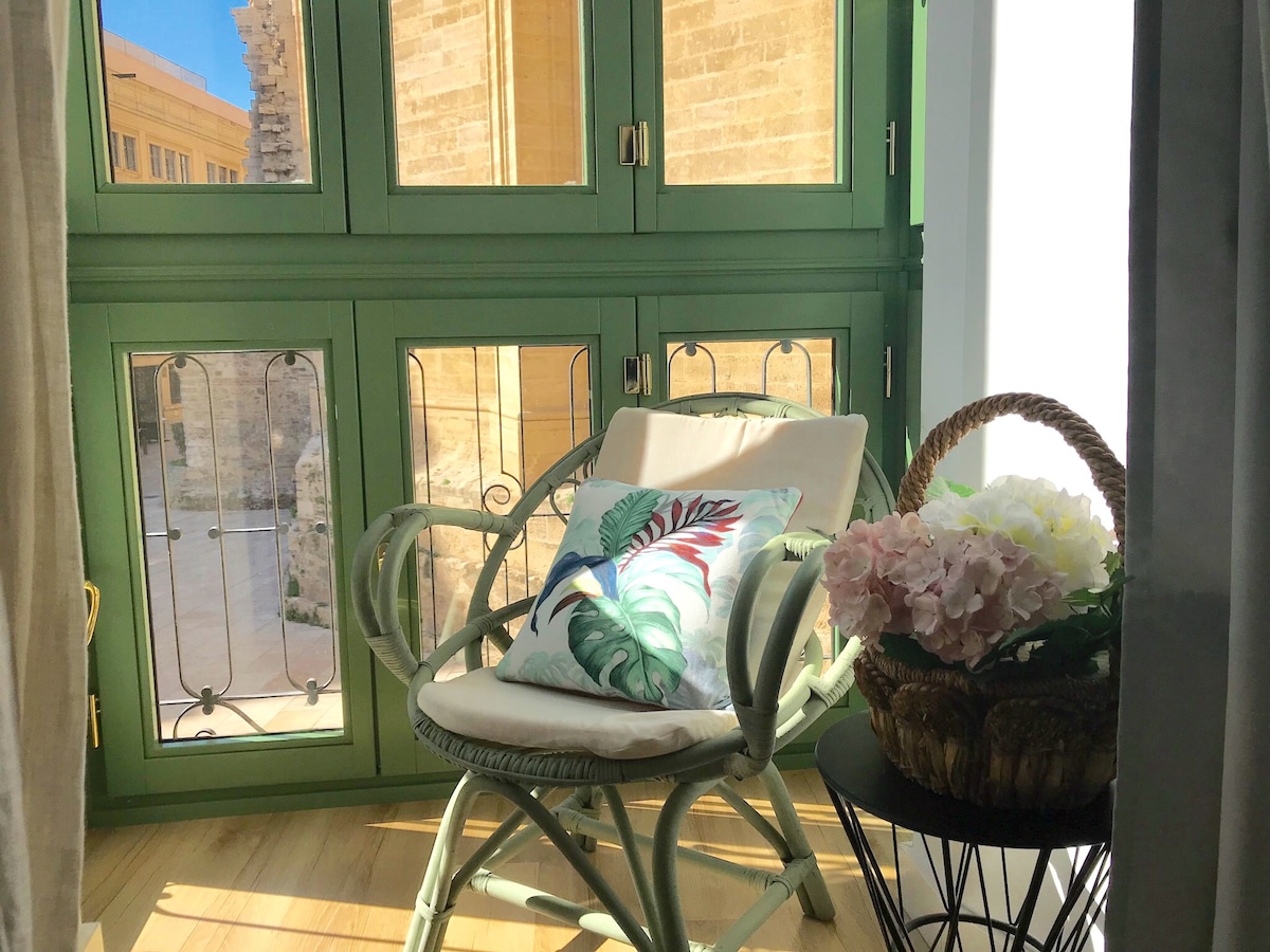 A light-filled corner features a comfortable chair adorned with a decorative pillow. A small side table is positioned next to the chair, and a basket holds flowers. The green-framed windows open to a view of the exterior, enhancing the space's bright atmosphere.
