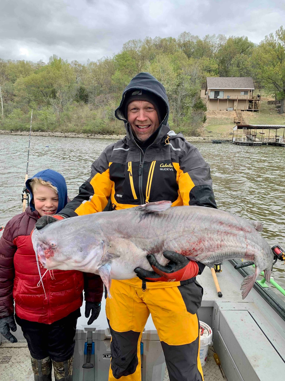 A person wearing a yellow and black waterproof suit holds a large catfish, while a child stands beside them, dressed in a red jacket. The scene is set on a boat with a lake in the background, displaying trees and a cabin on the shore.