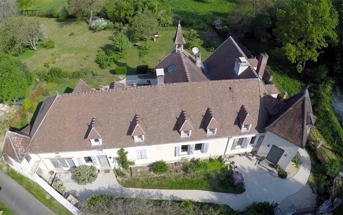 An aerial view of the historic hunting lodge from 1875 showcases its distinctive architecture with multiple gabled roofs. Surrounding green spaces and gardens are visible, providing ample outdoor areas. Pathways lead to various outdoor seating arrangements and leisure spots within the expansive property.