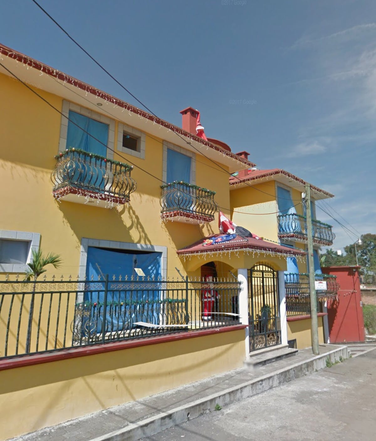 The exterior of a colorful two-story house is presented, featuring a bright yellow facade. Decorative iron balconies adorn the upper level, while a tiled roof and entry gate enhance the welcoming appearance. A landscaped area is visible in front, surrounded by fencing.