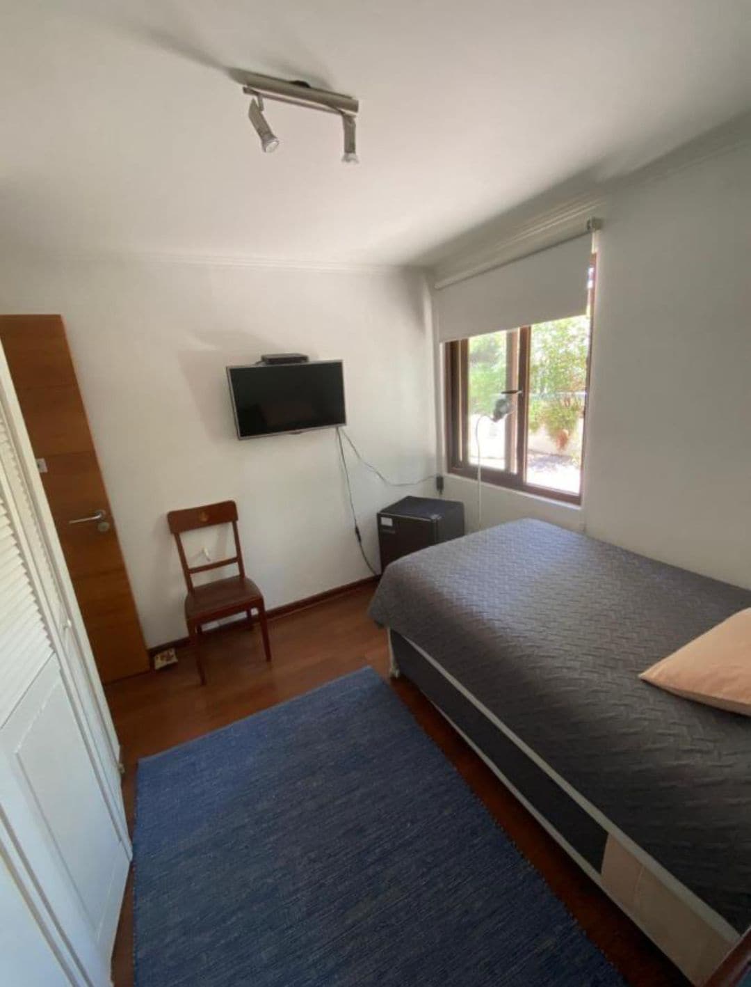 A simple bedroom is seen, featuring a single bed with light bedding positioned alongside a small desk and a chair. A flat-screen TV is mounted on the wall, and natural light filters in through the window, partially dressed with blinds. A small, dark rug covers the wooden floor.