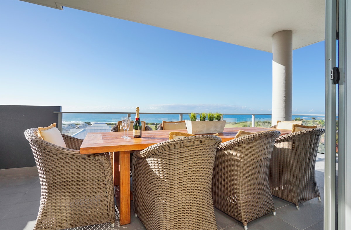 A spacious outdoor dining area features a large wooden table surrounded by eight wicker chairs. The setting offers panoramic ocean views, with sunlight illuminating the space, and is complemented by a potted plant in the center. The balcony provides a relaxing atmosphere for meals and gatherings.