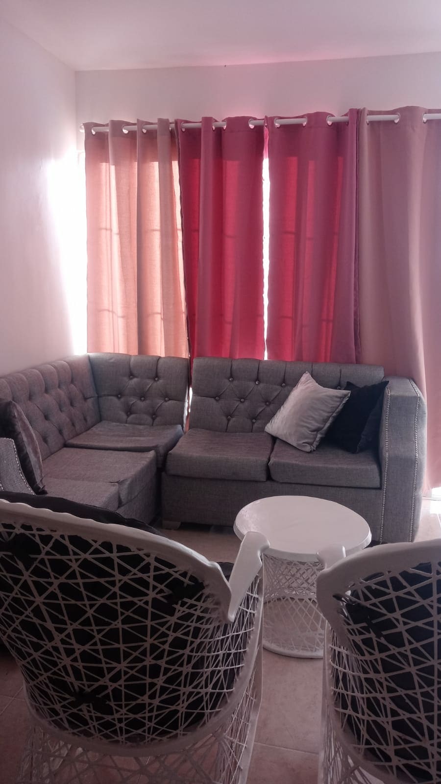 A cozy sitting area features a gray sectional sofa with tufted detailing, complemented by decorative pillows. Two white wicker chairs are positioned nearby, surrounding a small, circular table. Natural light streams through the curtain-covered windows, creating a warm ambiance.