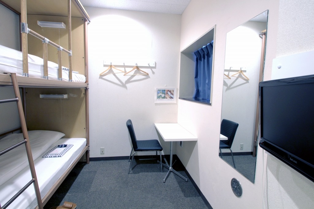 A cozy bunk bed room is presented with a wooden bunk bed and a comfortable chair positioned at a small table. A large mirror reflects the space, while hangers are attached to the wall. A window covered with blue curtains allows natural light to filter in.