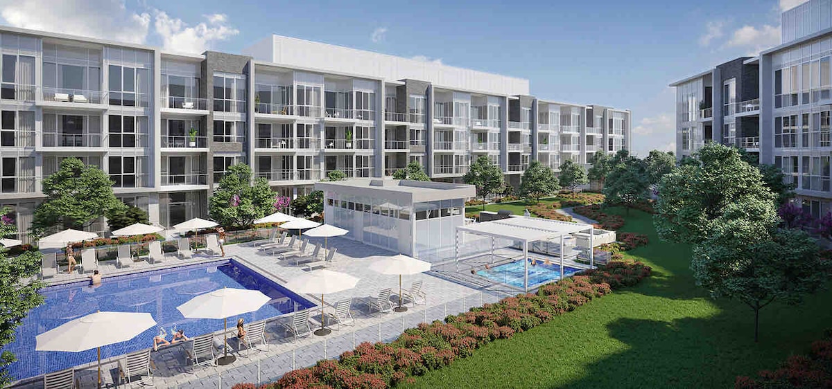 A spacious outdoor area is depicted, featuring a large pool surrounded by lounge chairs and umbrellas. Lush greenery and flowering plants line the edges, and modern multi-story buildings provide a backdrop. A shaded structure offers seating for relaxation near the water.