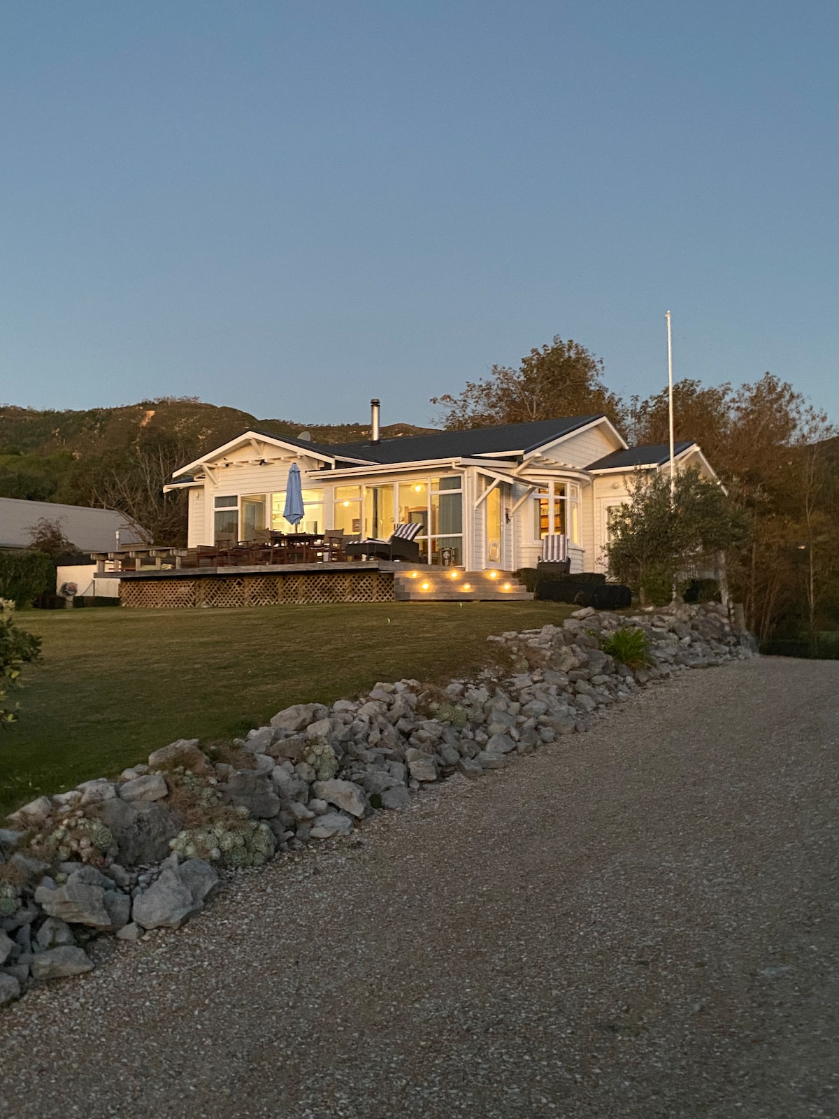 A charming house is positioned on a grassy area, gently illuminated by exterior lights. A spacious deck is visible, featuring outdoor seating and an umbrella. Surrounding landscaping includes well-kept shrubs and a rocky border, with hills in the background under a dusky sky.