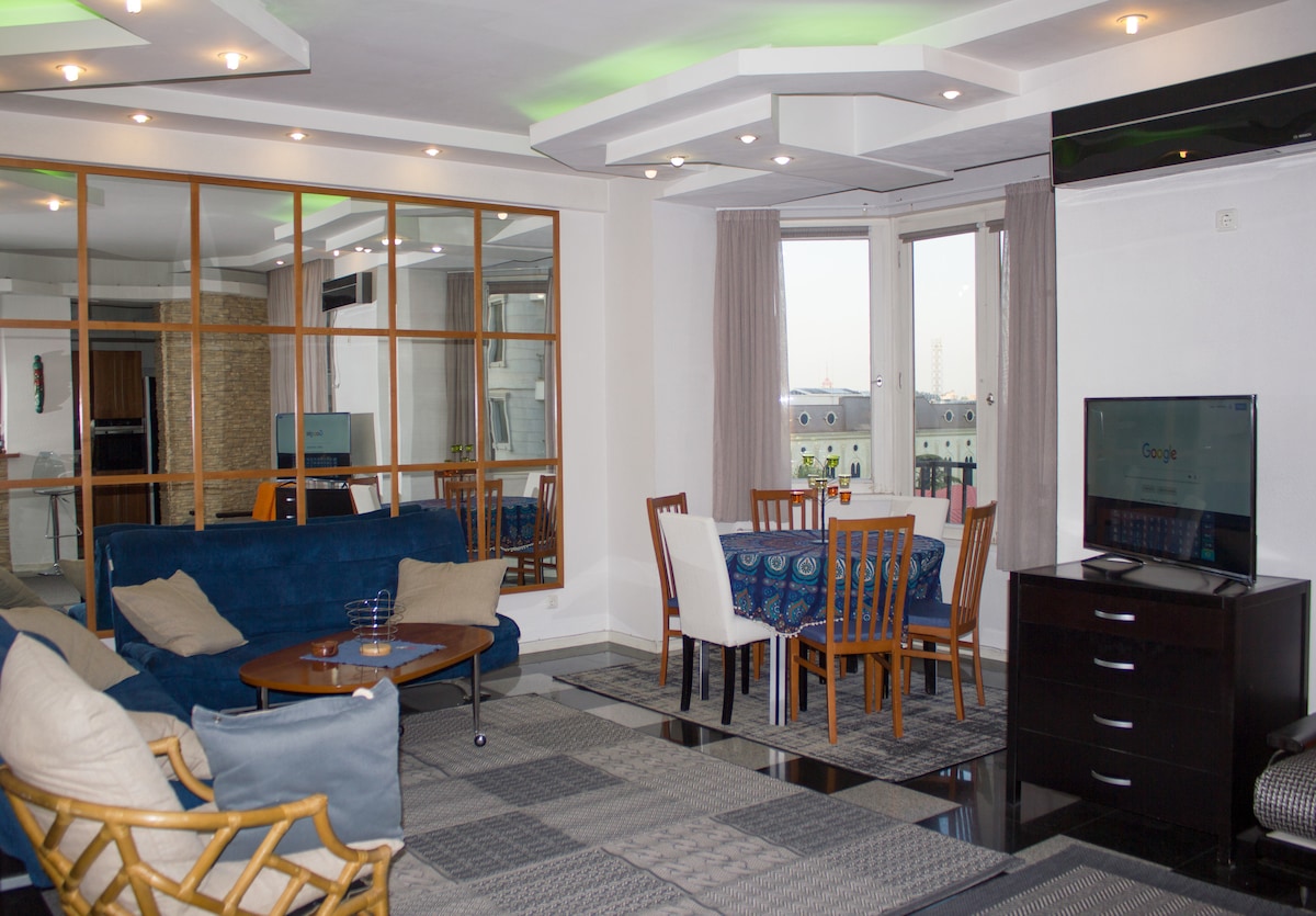 An open plan living and dining area features a round dining table with chairs and two convertible couches. A Smart TV is positioned next to a glass door leading to a balcony. The space includes large windows providing abundant natural light.