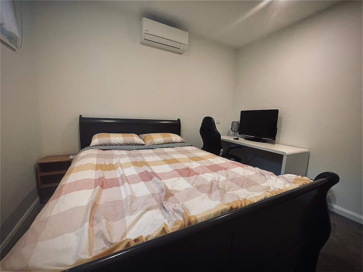 A cozy bedroom features a queen-sized bed with a plaid patterned duvet. An ergonomic chair is positioned at a desk next to a monitor. A ductless air conditioning unit is mounted on the wall, providing comfort throughout the year.