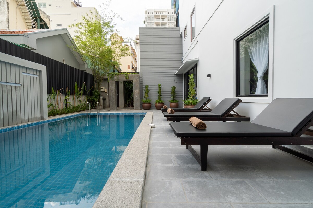 An outdoor area features a glistening swimming pool surrounded by a sleek stone patio. Two reclining lounge chairs are positioned beside the pool, each equipped with rolled towels. Lush greenery and modern architecture complete the serene environment.