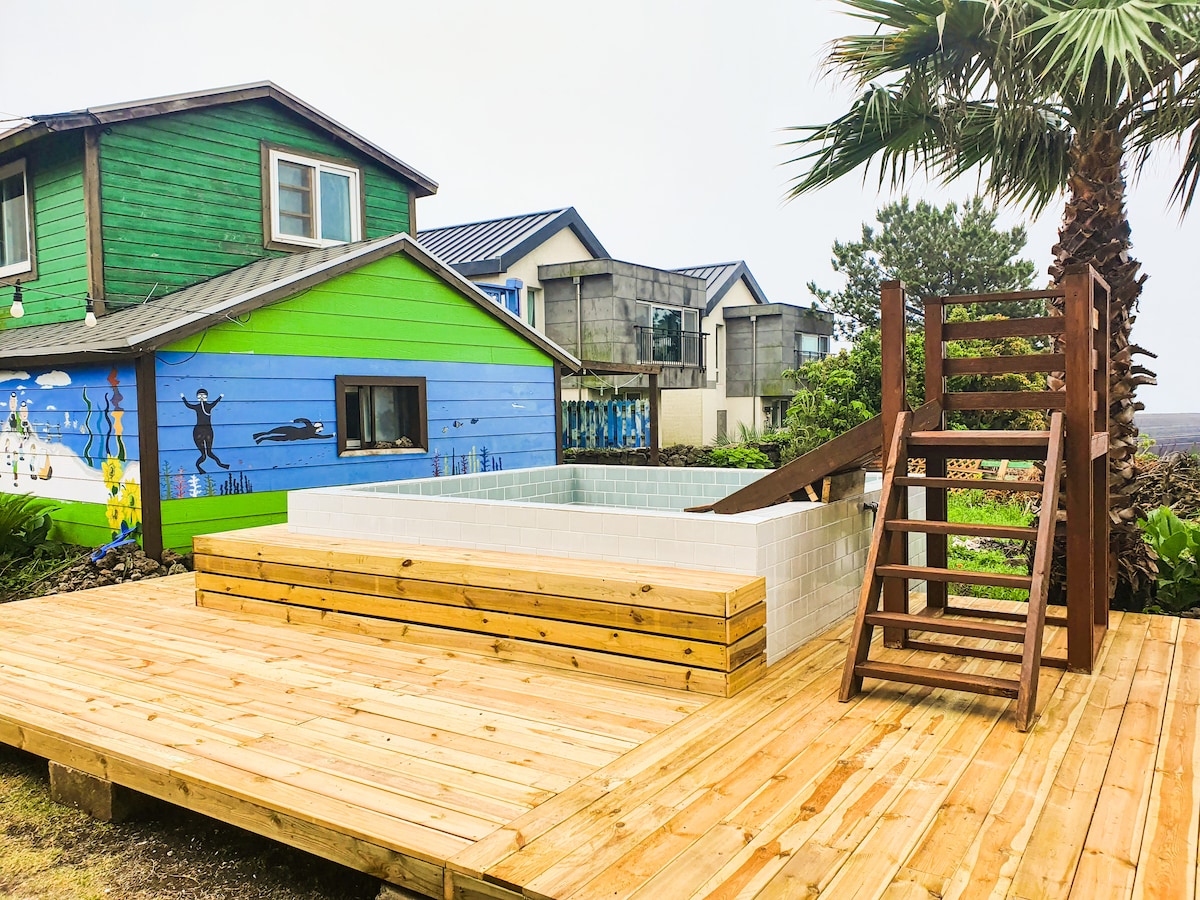 A wooden deck features a spacious hot tub, framed by palm trees and colorful murals on nearby buildings. A ladder provides access to the hot tub area, while the surrounding outdoor space is designed for relaxation and enjoyment.