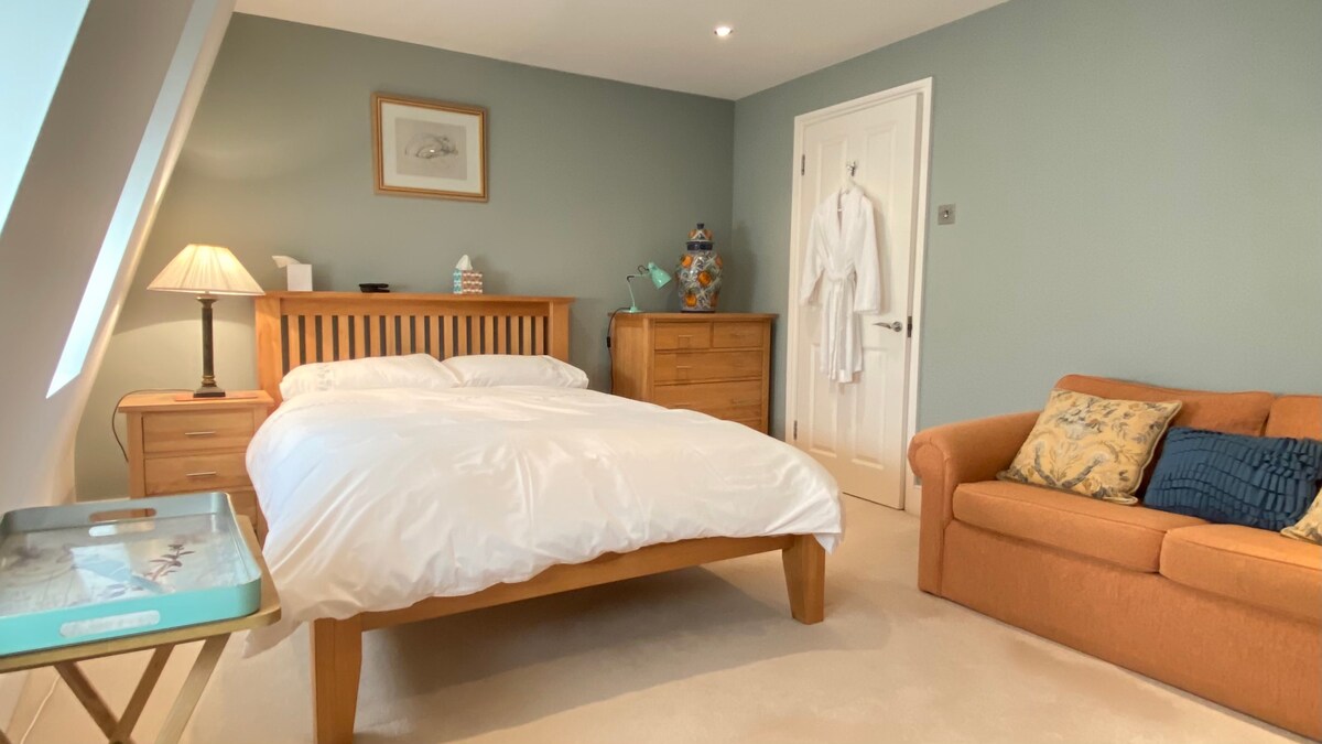 A bright and spacious room features a double bed with a white duvet, a wooden headboard, and light-colored bedside lamps. A cozy sofa and a dresser are present, along with a terracotta robe hanging on the door. Soft lighting enhances the serene atmosphere.