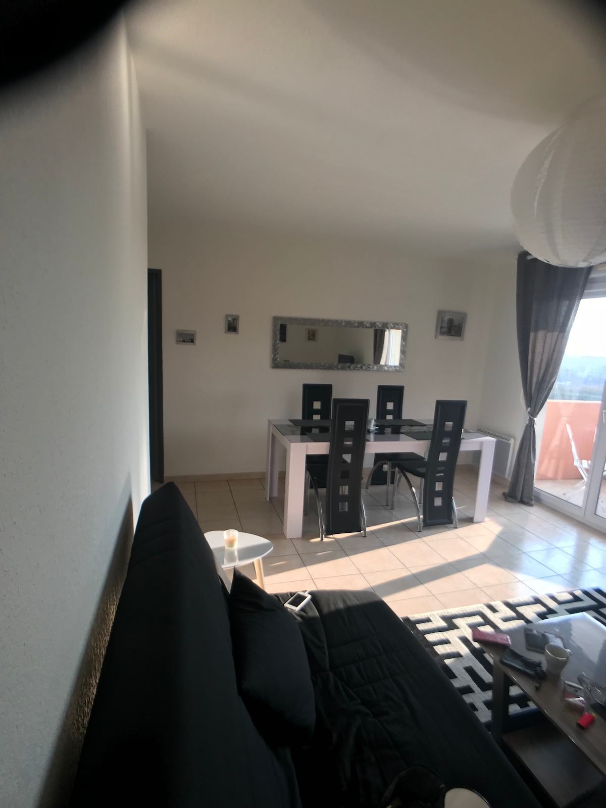 A modern dining area is visible, featuring a rectangular table surrounded by four black chairs. Soft natural light streams through large windows, illuminating the space. A black sofa and small side table are positioned nearby, adding functionality to the open layout.
