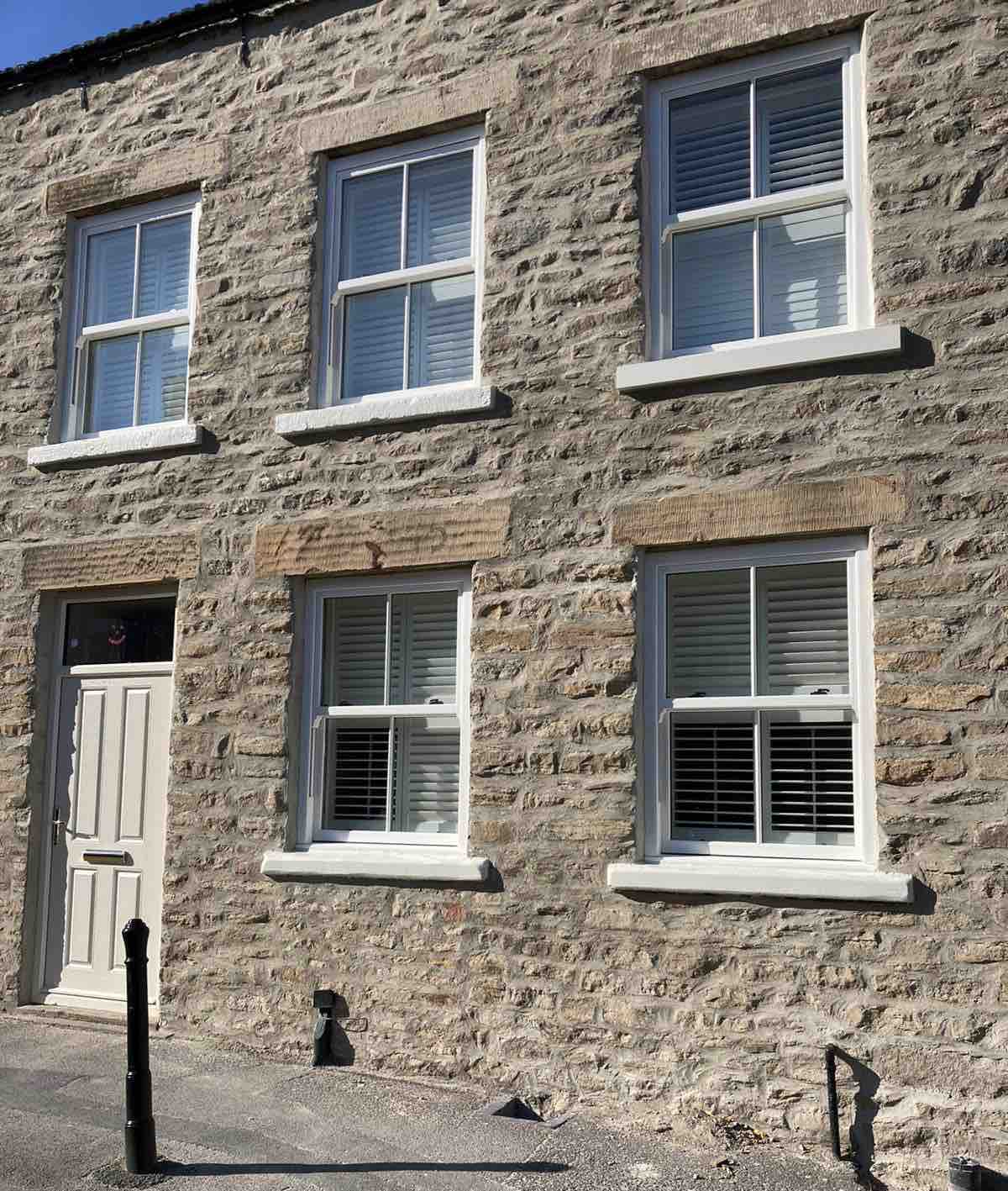 The front exterior of the cottage features a classic stone facade with three large windows that have white shutters. A solid front door adds to the welcoming appearance. The pavement is neatly finished, contributing to the cottage’s charming character.