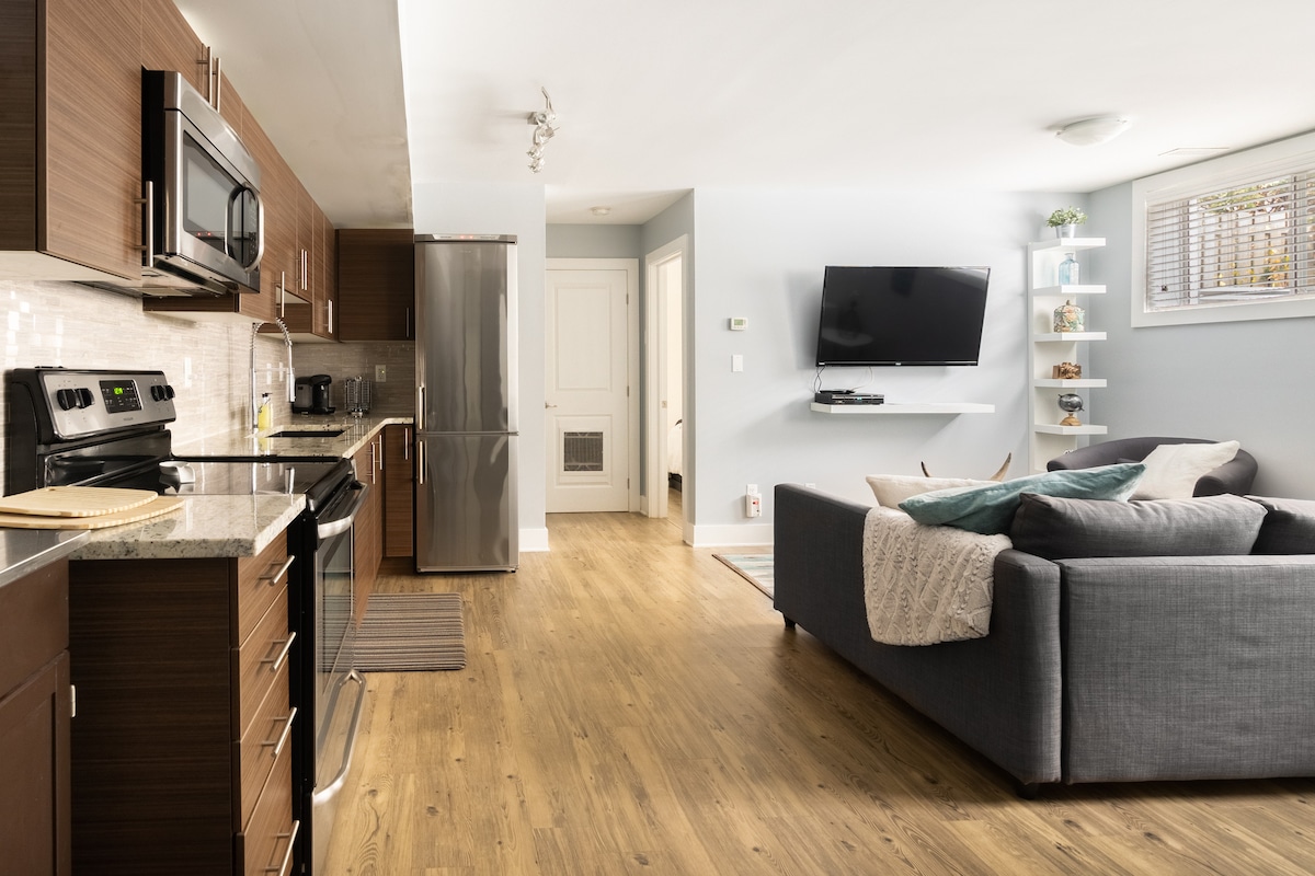 The kitchen and living area are shown in an open layout, featuring modern appliances, wooden cabinetry, and a spacious countertop. A comfortable gray sofa is positioned near a wall-mounted television, with natural light coming through a window adorned with blinds.