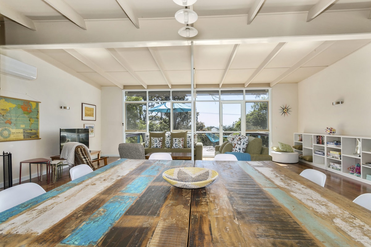 A spacious dining area features a large wooden table with a weathered finish, surrounded by white chairs. Natural light fills the room through expansive windows, showcasing a view of the outdoors. Comfortable seating is available in the corner, with a television and a bookshelf visible.