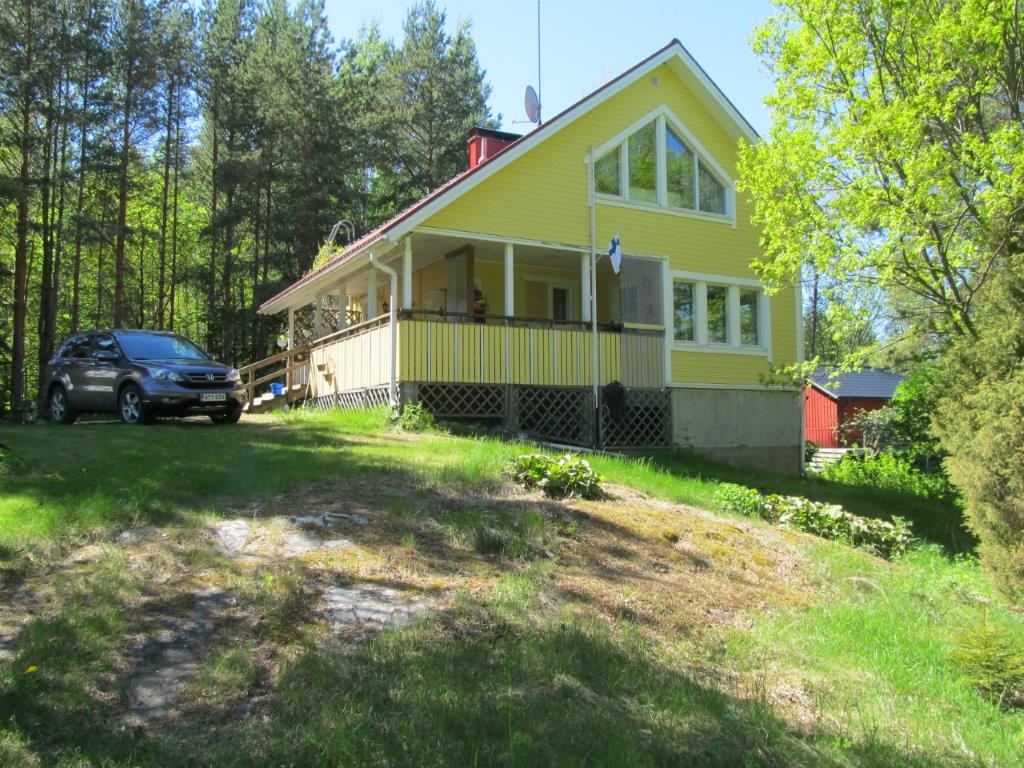 A yellow house with large windows is set amidst trees and green grass. The front porch features a railing, and a car is parked nearby. A stone pathway leads to the entrance, enhancing the natural surroundings.