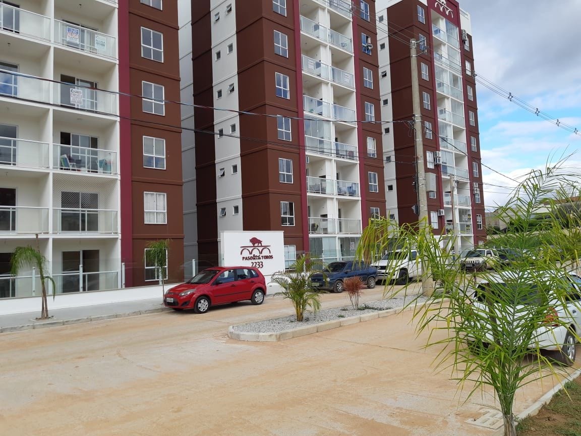A modern apartment building is shown, featuring multiple stories with a mix of red and brown facades. A parking area is visible in the foreground, accommodating various cars, while small palm trees are planted along the pathway.