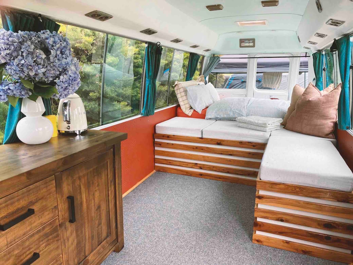 A cozy seating area is highlighted by large windows, allowing ample natural light to fill the space. Soft cushions and a throw pillow are arranged on a padded bench, while a wooden cabinet adds warmth and storage. Decorative flowers are positioned on the cabinet.