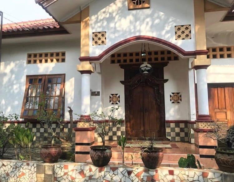 The front entrance is framed by decorative wooden doors, complemented by ornate carvings. Potted plants are arranged along the entryway, enhancing the welcoming vibe. Glass windows with grid patterns allow natural light to enter, while patterned tiles create a visually appealing ground area.