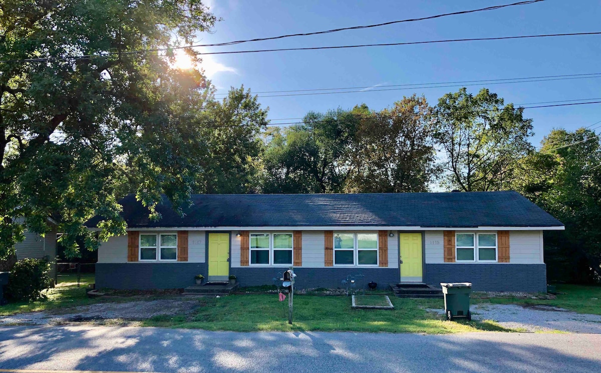 A well-maintained, two-unit structure is nestled among trees, displaying a gray exterior with wooden accents. Sunlight bathes the scene, highlighting the entryways marked by vibrant green doors. A grassy area is visible in the foreground, along with a mailbox and trash bin.