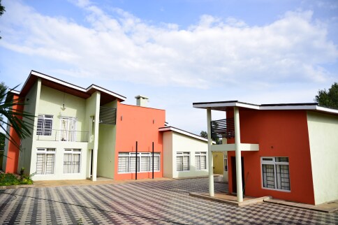 The exterior of Meru Forest House is presented, featuring a contemporary design with two main buildings. The vibrant colors include peach and light green. A patterned driveway enhances accessibility, while the expansive outdoor area is surrounded by lush greenery.