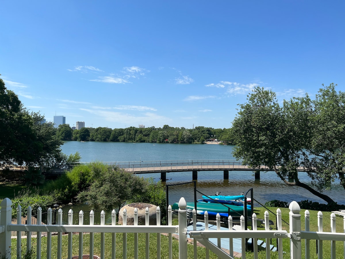 A scenic view of Lady Bird Lake is revealed, showcasing calm waters and lush greenery along the shoreline. A wooden bridge crosses the lake in the distance, while two kayaks are visible by the lakeside dock. A white picket fence frames the foreground.