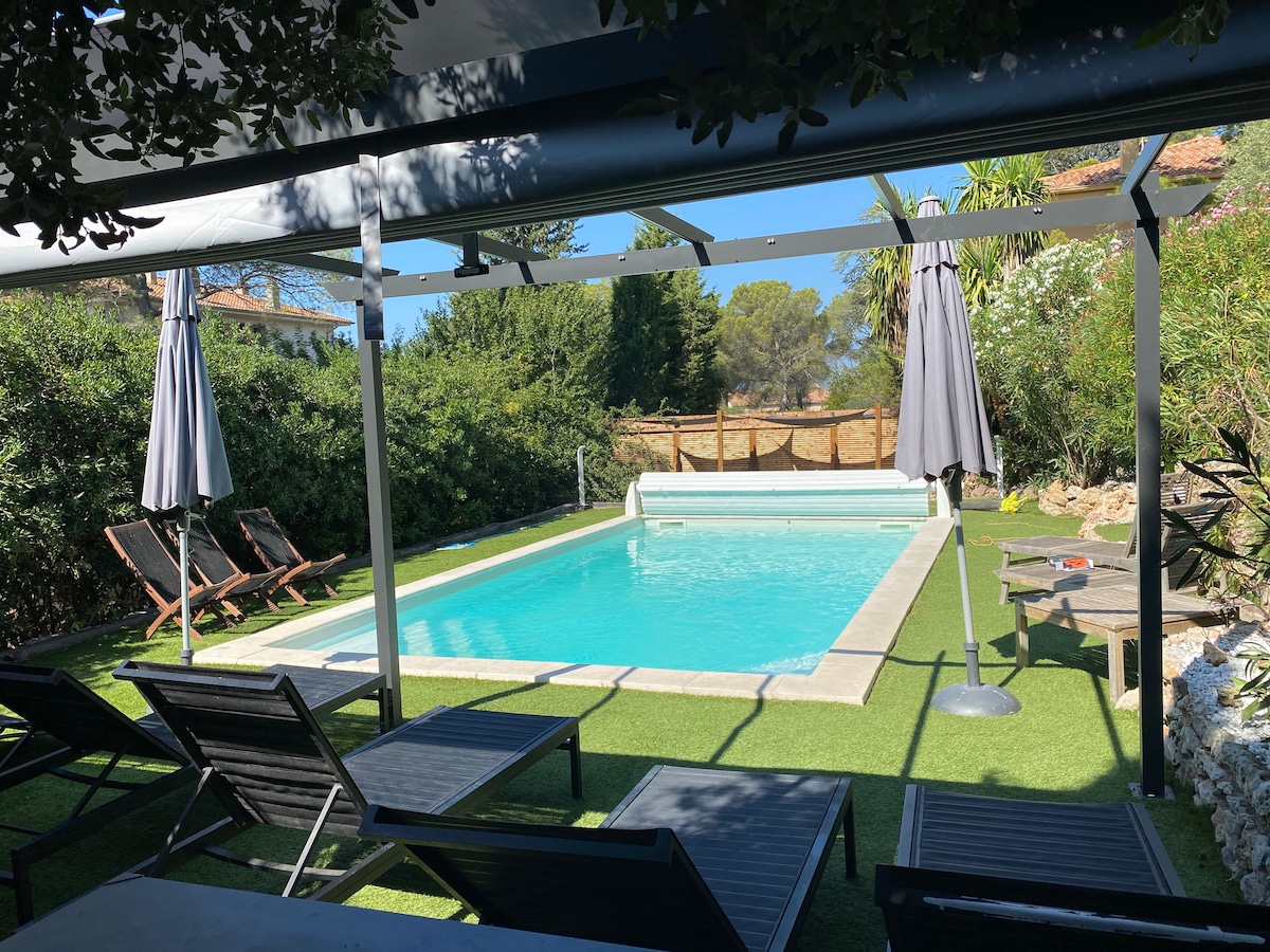 A serene pool area is presented, featuring a turquoise swimming pool bordered by a well-groomed lawn. Two sun loungers and two umbrellas provide shaded relaxation options. The surrounding greenery adds a natural feel, while a protective cover ensures comfort during warm days.
