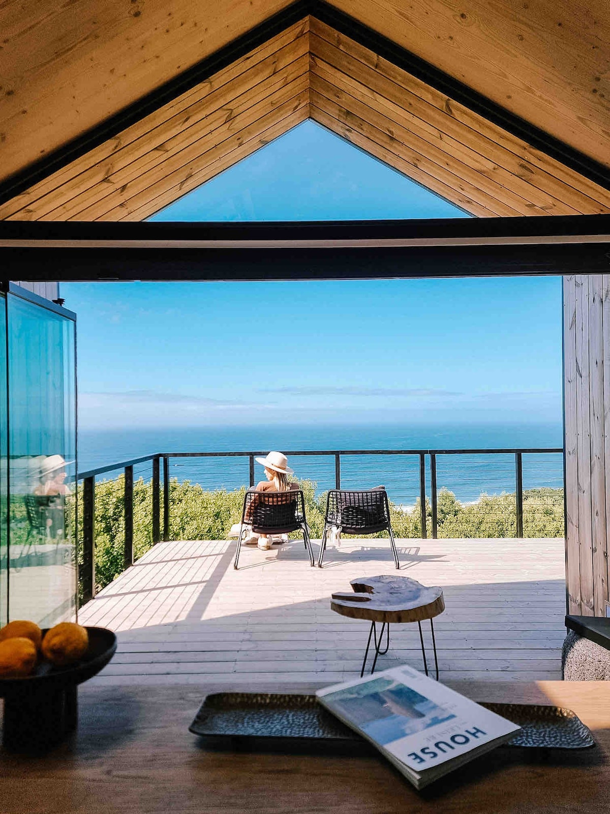 A stunning view from the cabin's interior showcases an expansive deck with two lounging chairs positioned against a backdrop of blue sea and sky. Sunlight floods the space, highlighting the wooden beams of the ceiling and the natural scenery outside.
